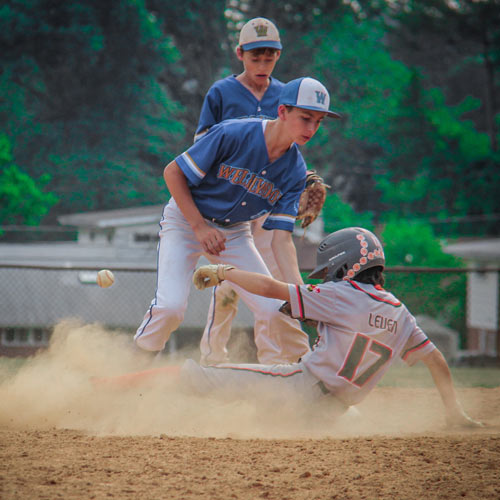 An Elkridge Hurricanes baserunner sliding into second base safely as the baseball thrown by the catcher ricochets off his helmet past the shortstop and into the outfield.